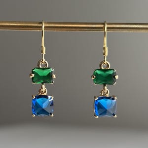 May include: Gold-toned dangle earrings with emerald green and sapphire blue rectangular gemstones. The earrings are displayed on a gold bar against a neutral gray background. The gemstones are faceted and catch the light.