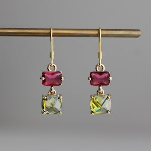 以下が含まれることがあります： ゴールドカラーのドロップイヤリングのペア。各イヤリングは、正方形の明るい緑色の宝石の上に、長方形のマゼンタ色の宝石が特徴です。イヤリングは、ゴールドカラーのバーに表示されています。