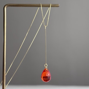 May include: A gold-toned box chain necklace featuring a long, vertical drop design that terminates in a single, translucent, teardrop-shaped pendant in a vibrant red color. The delicate metal chain is suspended from a thin, L-shaped metal stand.