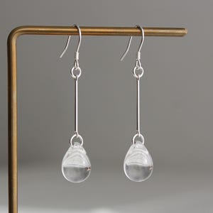 May include: A pair of silver drop earrings featuring a thin vertical metal bar suspended from a hook, ending in a clear, smooth, teardrop-shaped glass bead. The earrings are displayed on a thin, L-shaped metallic stand with a polished finish.