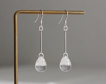 Könnte beinhalten: Ein Paar silberne Ohrhänger mit einem dünnen, vertikalen Metallstab, der an einem Haken befestigt ist und in einer klaren, glatten, tropfenförmigen Glasperle endet. Die Ohrringe sind an einem dünnen, L-förmigen Metallständer befestigt.