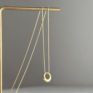 Puede incluir: Un collar de tono dorado que presenta una cadena fina de eslabones cuadrados y un pequeño colgante circular liso. El colgante tiene una superficie pulida y reflectante con un perfil redondeado y grueso. El collar cuelga de un soporte metálico.