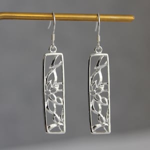 May include: A pair of silver-toned drop earrings featuring a rectangular frame with an intricate, openwork floral cutout design. Each earring is suspended from a simple silver-toned hook, showcasing a polished metallic finish and a slender, elongated silhouette.