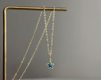 May include: A gold-toned chain necklace featuring a small, five-pointed star pendant. The star pendant has a dark blue, speckled, and iridescent inlay set within a thin gold-colored metal border, suspended from a delicate link chain.