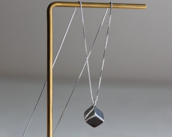 May include: A silver necklace with a geometric cube pendant. The pendant is suspended from a delicate chain and hangs from a gold-colored display stand. The cube has a dark, textured surface with subtle highlights.