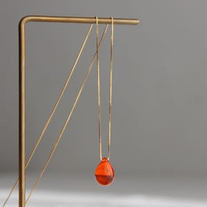 May include: A gold chain necklace with a teardrop-shaped pendant made of orange glass.