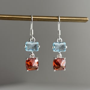 May include: A pair of drop earrings featuring silver hooks, each with a rectangular light blue gemstone suspended above a square, faceted deep orange-red gemstone. The stones are held in place by silver prongs, creating a dangling design.