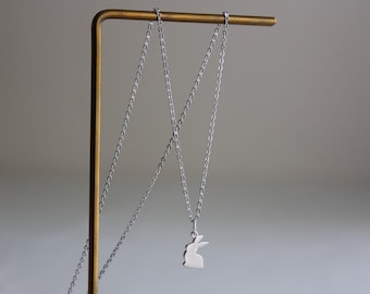 May include: A silver chain necklace with a silver rabbit pendant hanging from it. The necklace is displayed on a gold metal stand.