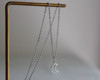 May include: A silver chain necklace with a crescent moon pendant. The necklace is hanging on a gold metal display stand.