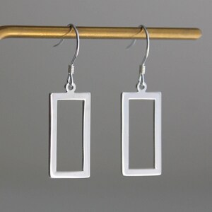 May include: A pair of silver-toned earrings featuring a hollow rectangular frame design. Each earring is suspended from a curved metal hook, showcasing a clean, open-centre geometric shape with a smooth, polished metallic finish throughout.