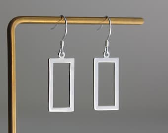 May include: A pair of silver-toned earrings featuring a hollow rectangular frame design. Each earring is suspended from a curved metal hook, showcasing a clean, open-center geometric shape with a smooth, polished metallic finish throughout.