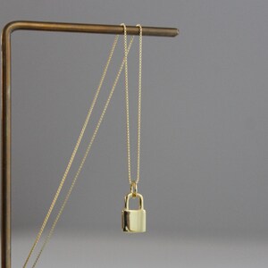 May include: A gold-colored padlock pendant necklace. The pendant is a small, rectangular padlock with a rounded top. The necklace chain is delicate and gold-colored. The necklace is displayed on a gold-colored stand against a gray background.