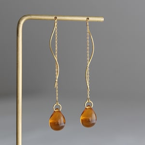 Op de afbeelding: Een paar gouden oorbellen met een traanvormige amberkleurige steen die aan elke oorbel hangt.