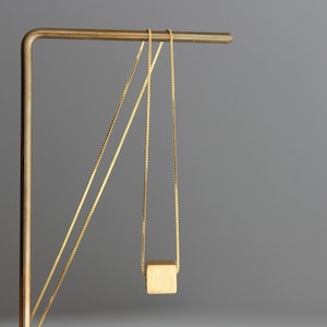 May include: A gold-coloured necklace featuring a thin box chain and a small, solid cube pendant with a matte finish. The chain is draped over a thin, L-shaped metal stand, allowing the cube to hang freely from the centre of the delicate chain.