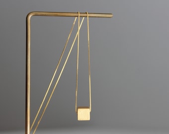 May include: A gold-toned necklace featuring a delicate box chain and a single, small, solid cube pendant. The pendant has a matte, textured finish and hangs centered from the thin, metallic chain, which is draped over a gold-colored stand.