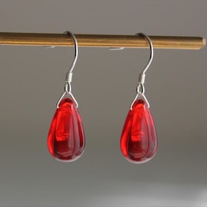 May include: A pair of drop earrings featuring translucent red teardrop-shaped pendants suspended from silver-toned metal hooks. The smooth, polished red glass elements catch the light, creating a clear, vibrant appearance for these dangling earrings.