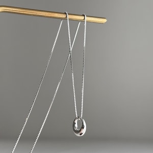 May include: A silver-toned necklace featuring a delicate box chain and a small, smooth, circular pendant with a polished, reflective finish. The necklace is draped over a thin, L-shaped brass-coloured metal stand, showcasing the chain's length.