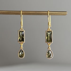 May include: A pair of gold-toned drop earrings featuring a rectangular olive green gemstone set above a smaller, pear-shaped olive green gemstone with a beaded edge. The earrings hang from simple gold-toned hooks, showcasing a symmetrical design.