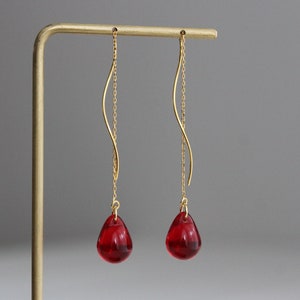 Pode incluir: Um par de brincos de argola dourados com contas de vidro vermelhas em forma de lágrima. Os brincos têm uma corrente delicada e um design de fio curvo.