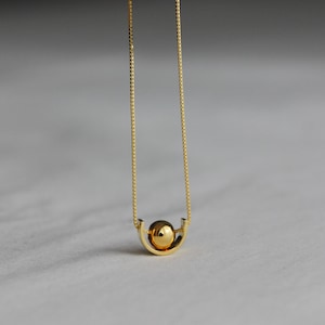 Peut inclure: Un collier doré composé d'une fine chaîne à maillons carrés et d'un pendentif formé d'une sphère lisse et polie reposant dans un berceau métallique incurvé en forme de U. L'ensemble présente une finition métallique réfléchissante.