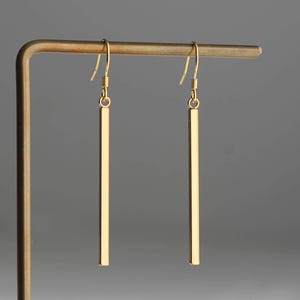 May include: A pair of gold-toned drop earrings featuring long, slender, rectangular bar pendants suspended from simple curved ear wires. The earrings have a smooth, polished metallic finish and hang vertically from a thin, L-shaped metal display stand.