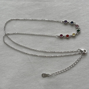 Op de afbeelding: Een zilverkleurige ketting met een reeks van zeven kleine, ronde, veelkleurige edelstenen op een rij. De ketting is voorzien van een fijne schakelketting, een karabijnsluiting en een verstelbare verlengketting met een klein metalen label.