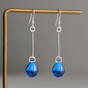 Peut inclure: Une paire de boucles d'oreilles pendantes composées d'un crochet argenté, d'une tige verticale droite et d'une perle en verre translucide bleu profond en forme de goutte. Les boucles sont suspendues par un anneau en fil d'argent fin.
