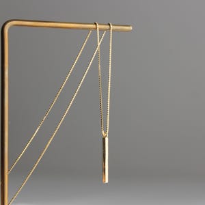 May include: A gold necklace with a long, thin, rectangular pendant hanging from it. The necklace is displayed on a gold metal stand.
