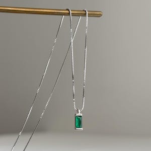 May include: A silver necklace featuring a rectangular, emerald-green pendant. The pendant is set in a silver frame and suspended from a fine silver chain. The necklace is displayed on a gold-coloured jewellery stand against a neutral backdrop.