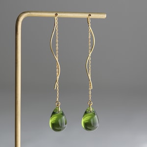 Może przedstawiać: Para złotych kolczyków. Każdy kolczyk ma delikatny łańcuszek, który wdzięcznie się zakrzywia, zawieszając przezroczysty, zielony kamień szlachetny w kształcie łzy. Kolczyki są prezentowane na neutralnym szarym tle.