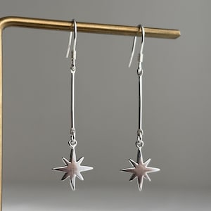 May include: A pair of silver-colored dangle earrings. Each earring features a long, thin bar suspended from a hook, with a star-shaped charm at the bottom. The earrings are displayed against a neutral gray background.