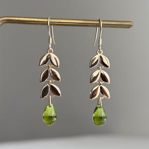 Op de afbeelding: Goudkleurige oorhangers met een bladvormgeving. Elke oorbel heeft drie bladvormige elementen en een druppelvormige groene edelsteen aan de onderkant. De oorbellen worden getoond op een goudkleurige sieradenstandaard.