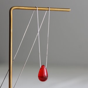 May include: A necklace featuring a small, smooth, translucent red teardrop-shaped pendant suspended from a thin, silver-coloured box chain. The pendant has a glossy finish and hangs vertically, showcasing a vibrant crimson hue against the metal chain.
