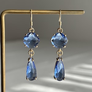 Peut inclure: Une paire de boucles d'oreilles pendantes avec des cristaux bleus à facettes. Chaque boucle se compose d'un cristal carré en haut relié à un cristal en forme de goutte en bas, montés sur des griffes en métal doré et suspendus à des crochets dorés.