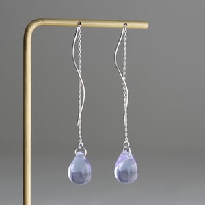 May include: A pair of silver drop earrings with a delicate, curved design. Each earring features a slender chain and a teardrop-shaped, lavender-colored gemstone. The earrings are displayed against a neutral gray backdrop.