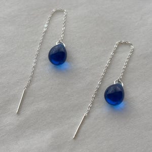 May include: A pair of dangle earrings with a silver chain and a teardrop-shaped, deep blue glass bead. The earrings have a minimalist design, with a straight silver bar at the end of the chain.