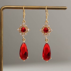 May include: Gold dangle earrings featuring a red teardrop-shaped gemstone and a square-shaped gemstone with a filigree design.