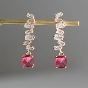 Peut inclure: Une paire de boucles d'oreilles en or avec une pierre précieuse carrée rouge pendante et une rangée de pierres de forme baguette transparentes.