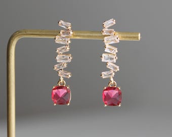 May include: A pair of gold-tone earrings featuring a vertical row of clear baguette-cut cubic zirconia stones arranged in a staggered pattern, terminating in a single, square-cut, deep pink gemstone drop suspended from the base of each earring.