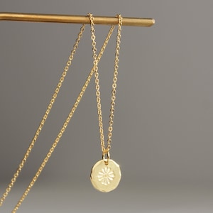 May include: A gold-coloured necklace with a small, round pendant featuring a floral design. The pendant hangs from a fine chain. The necklace is displayed on a gold-coloured stand against a grey background.