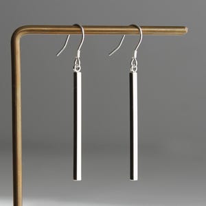 May include: A pair of silver-toned drop earrings featuring a long, slender, polished cylindrical bar pendant suspended from a simple curved ear wire hook. The metal surface reflects light, creating a bright, smooth, and metallic appearance.