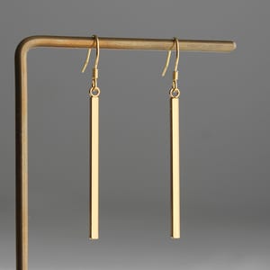 Op de afbeelding: Een paar gouden staafjes oorbellen die aan een displaystandaard hangen. De oorbellen zijn lang en dun, met een eenvoudig, minimalistisch ontwerp.