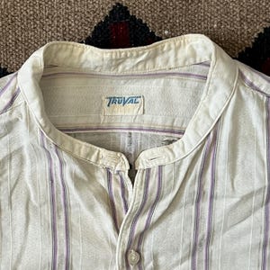Op de afbeelding: Crèmekleurig overhemd met verticale paarse strepen. Het overhemd heeft een bandkraag en een klein label met de tekst "TRUVAL" in een blauw en wit ontwerp. De stof lijkt licht van gewicht.