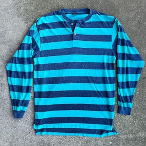 May include: A long-sleeved striped shirt with a blue and turquoise color scheme. The shirt has a button-up collar and a single button on the front.
