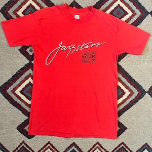 May include: A red t-shirt with the text "Milestone Jazzstars American Tour Fall 78". The text is in a stylized font. The shirt has a crew neck and short sleeves. The graphic is in white and black.