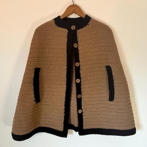 Op de afbeelding: Een bruin gehaakte cape met zwarte randen en knopen. De cape heeft een ronde halslijn, twee voorzakken en een knoopsluiting. De zwarte randen omlijnen de randen, zakken en knooplijst.