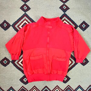 May include: A red zip-up jacket with a ribbed texture on the upper portion and two large pockets on the lower portion.