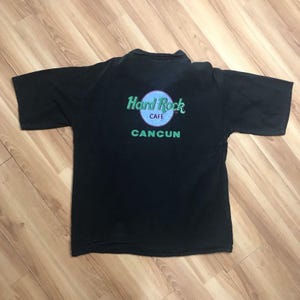 Könnte beinhalten: Schwarzes T-Shirt mit kurzen Ärmeln und einem grünen Hard Rock Cafe Logo sowie dem Aufdruck "CANCUN" auf der Rückseite.