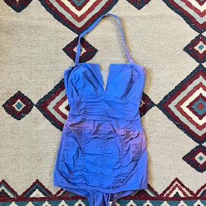 May include: A vintage blue one-piece swimsuit with a halter neck and a V-neckline. The swimsuit features ruched detailing on the bodice and a classic silhouette. The fabric has a slight ombre effect, transitioning from a lighter to a darker shade of blue.