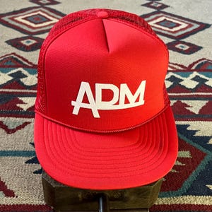 Peut inclure: Casquette de camionneur rouge vif avec le logo "ADM" blanc. Le chapeau a un dos en filet et une visière incurvée. Le chapeau est posé sur une surface en bois. L'arrière-plan est un tapis à motifs.